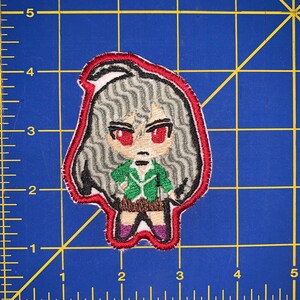 Puede incluir: Parche bordado de un personaje de dibujos animados con cabello largo gris, ojos rojos y una chaqueta verde. El personaje lleva una falda marrón y zapatos morados. El parche tiene un contorno rojo.