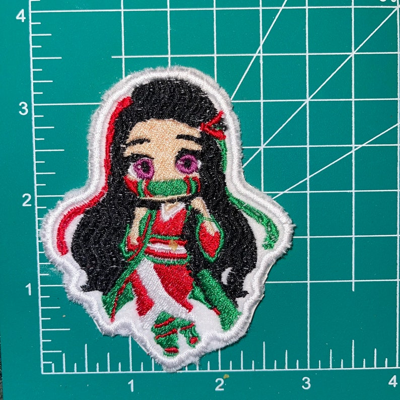 May include: An embroidered patch featuring a cartoon character with black hair, a green and red outfit, and a green mask. The character is wearing a white and red patterned outfit and has purple eyes.