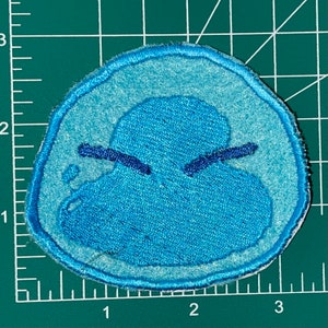 May include: A blue embroidered patch of a cartoon character with a frowning expression. The character is a blue blob with two small eyes and a mouth.