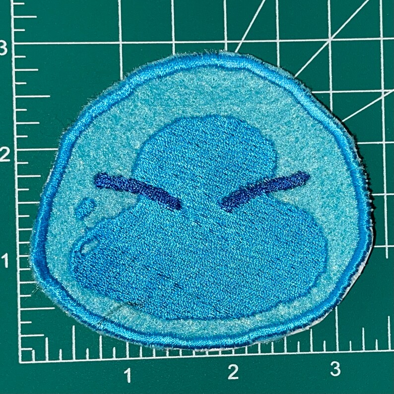 May include: A blue embroidered patch of a cartoon character with a frowning expression. The character is a blue blob with two small eyes and a mouth.