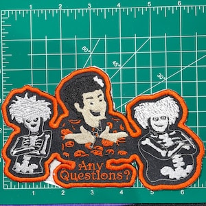 May include: An orange embroidered patch with a cartoon character in the center. The character is wearing a black shirt with a white skull and crossbones pattern. The character is surrounded by two skeletons with white bones and black bodies. The text "Any Questions?" is below the character.