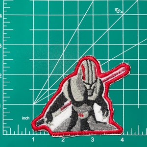 May include: Red and gray embroidered patch of a character holding a red sword. The character is wearing a gray suit with a red belt and has a red stripe on its head.