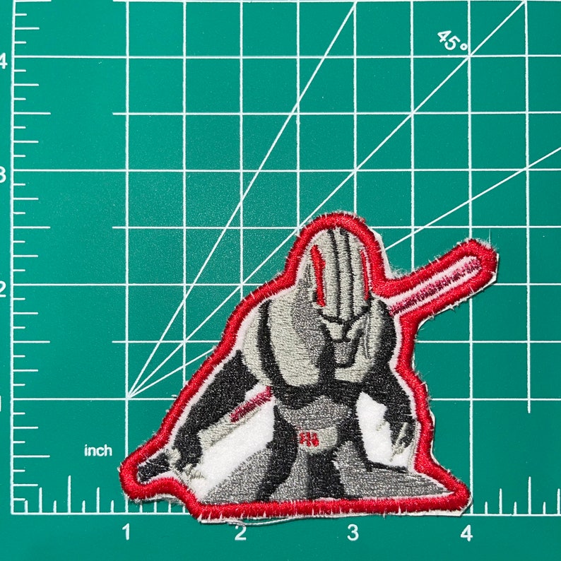 Op de afbeelding: Rode en grijze geborduurde patch van een personage dat een rood zwaard vasthoudt. Het personage draagt een grijs pak met een rode riem en heeft een rode streep op zijn hoofd.