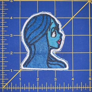 May include: Embroidered patch featuring a side profile of a blue-skinned woman with blue hair and pink lips. The patch has a white border and is on a blue grid background. The design is reminiscent of a cartoon or anime character.
