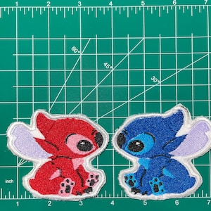 May include: Two embroidered patches of Stitch, a character from the Disney movie Lilo & Stitch. One patch is red and the other is blue. Both patches have white outlines and are sitting on a green grid.