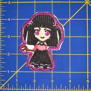 May include: Embroidered patch featuring a cartoon girl with black hair, purple eyes, and a black dress. The patch has a white background with a purple border. The girl is holding a brown object. The patch is approximately 3 inches tall.