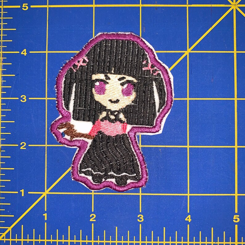 May include: Embroidered patch featuring a cartoon girl with black hair, purple eyes, and a black dress. The patch has a white background with a purple border. The girl is holding a brown object. The patch is approximately 3 inches tall.