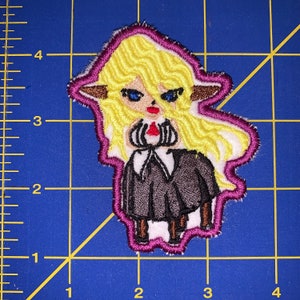 May include: Embroidered patch of a cartoon character with long blonde hair, wearing a black dress and red lipstick. The character has large blue eyes and brown ears. The patch is outlined in pink.