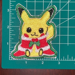 May include: A yellow Pikachu wearing a red and green Santa Claus costume with a white trim. The embroidered patch is on a green grid background.