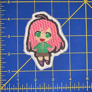 Puede incluir: Parche bordado con un personaje de estilo anime chibi con cabello rosado, ojos verdes y una chaqueta verde. El personaje lleva una falda marrón y botas moradas. El parche tiene un borde blanco y un contorno negro.