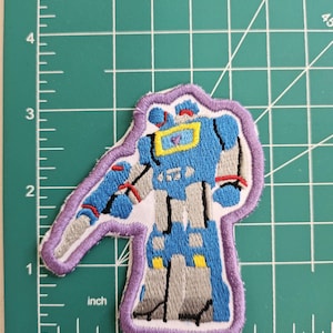 Op de afbeelding: Een geborduurde strijk-op patch met een blauwe robot met gele accenten. De robot staat met zijn armen uitgestrekt. De patch is omrand met paars.