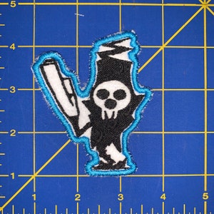 May include: A black and white embroidered patch of a skeleton holding a scythe, outlined in blue. The skeleton is wearing a hat and has a skull face.