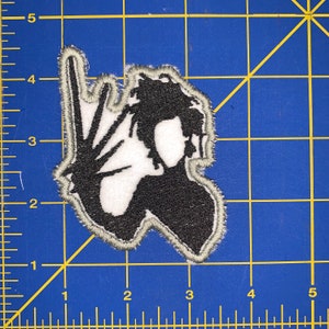 May include: Black and white embroidered iron-on patch of a silhouette of a person with long hair and a hand raised in a rock and roll hand gesture.