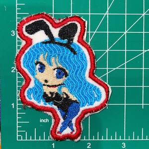 Pode incluir: Etiqueta bordada com um personagem de desenho animado com cabelo azul, vestindo um traje de coelho preto e um contorno vermelho.