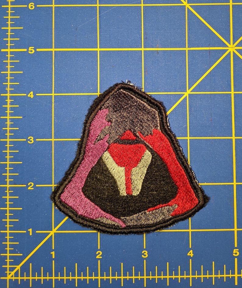 May include: A dark brown, pink, and red embroidered patch in the shape of a hooded figure. The figure is wearing a dark brown hooded cloak with a red lining. The hood is pulled up over the figure's head, and the figure's face is not visible.