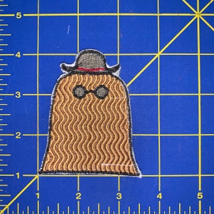 May include: A brown and tan embroidered patch of a cartoon character wearing a black hat and glasses. The character has a wavy, striped pattern.