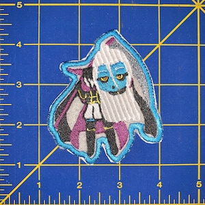 May include: An embroidered patch featuring a cartoon character with blue skin, white hair, and a purple and black outfit. The character is wearing a black cape and has a white and black striped pattern on their clothing.