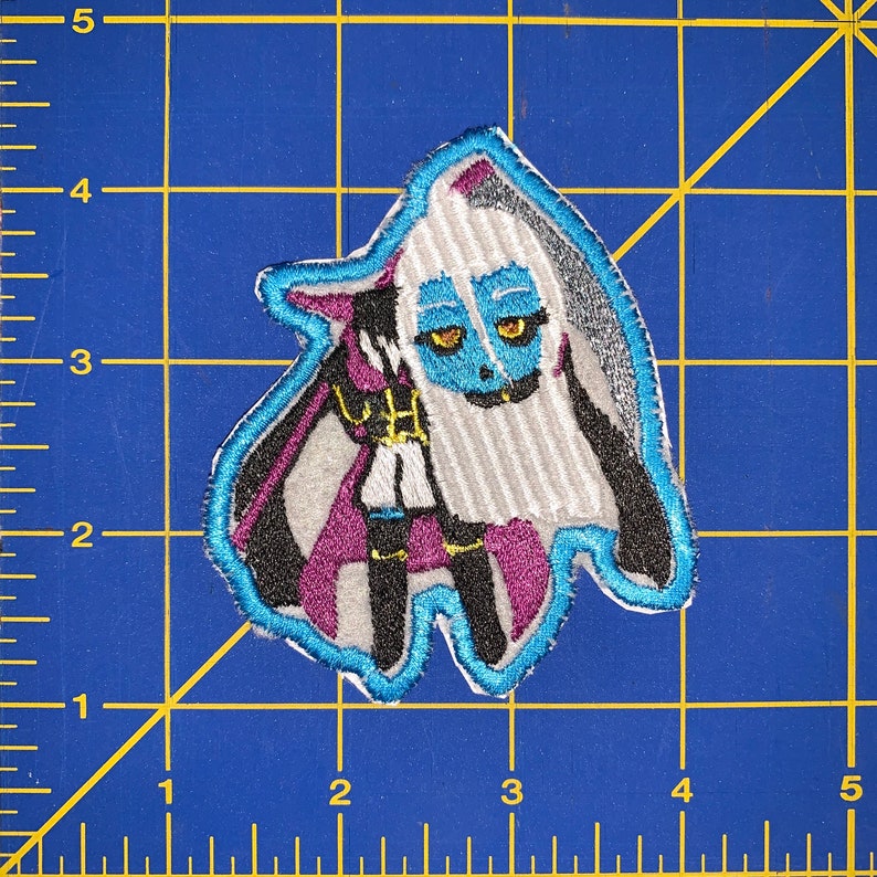 May include: An embroidered patch featuring a cartoon character with blue skin, white hair, and a purple and black outfit. The character is wearing a black cape and has a white and black striped pattern on their clothing.