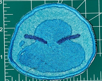 以下が含まれることがあります： しかめっ面をした漫画のキャラクターの青い刺繍パッチ。キャラクターは、小さな目が2つと口のある青い塊です。