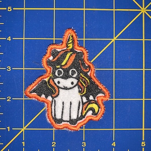 May include: An embroidered patch of a black and white unicorn with a yellow horn and mane. The unicorn has orange wings and is outlined in orange.