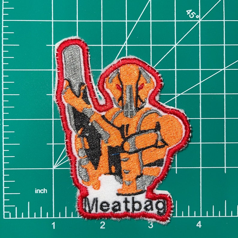 May include: Orange and gray embroidered patch with a robot-like figure holding a weapon. The patch has a red border and the text "Meatbag" in white letters.