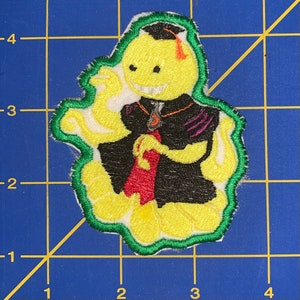 May include: Embroidered patch featuring a yellow character with a graduation cap and a red tie. The character is wearing a black robe with purple accents and a green border. The patch is on a blue grid.
