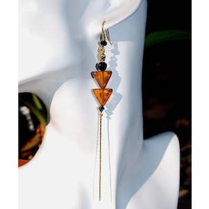 Peut inclure: Boucle d'oreille pendante unique avec un crochet doré, des perles noires et deux perles triangulaires ambrées. Une fine chaîne dorée pend du bas. La boucle d'oreille est présentée sur une tête de mannequin blanche.
