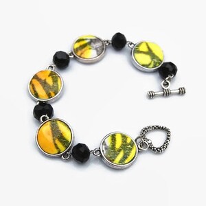 Peut inclure: Un bracelet argenté orné de breloques circulaires à motifs jaunes et noirs. Le bracelet est agrémenté de perles noires et d'un fermoir à bascule. Le design a une esthétique inspirée de la nature.