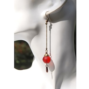 Peut inclure: Une seule boucle d'oreille longue avec une perle de verre rouge et un petit charme en forme de goutte d'eau rouge foncé. La boucle d'oreille est suspendue à un crochet et une chaîne de couleur or, présentée sur une tête de mannequin blanche. L'arrière-plan est flou avec des tons verts et bruns.