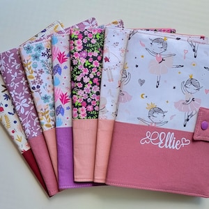 Peut inclure: Un ensemble de huit pochettes de livres en tissu avec des motifs floraux et de ballerines. Les pochettes sont roses, violettes et blanches. La plus grande pochette a un rabat rose avec le nom "Ellie" brodé dessus.