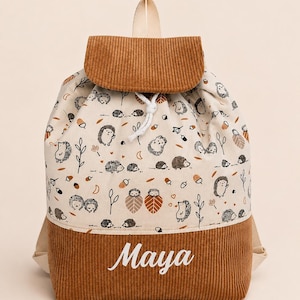Peut inclure: Un sac à dos à cordon en tissu crème avec un motif répétitif de hérissons, de feuilles et de glands. Le sac possède un rabat et une base en velours côtelé marron, avec le prénom Maya brodé en écriture blanche sur la poche avant.