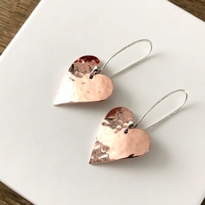 May include: A pair of hammered copper heart earrings with silver hooks. The earrings are hanging on a white surface.