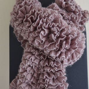 Peut inclure: Écharpe mauve avec un design à volants et une bordure blanche. L'écharpe est présentée sur un support noir, mettant en valeur sa texture et son style. Cet accessoire ajoute une touche d'élégance.