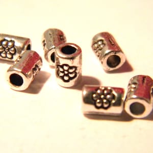 Puede incluir: Seis tapones de cuentas de metal plateado con un diseño floral. Los tapones de cuentas son cilíndricos y tienen un agujero en el centro.