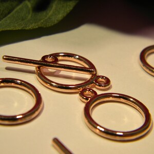 Pode incluir: Fecho de correr de metal dourado rosa com dois anéis redondos. O fecho de correr tem uma barra que desliza através dos anéis.