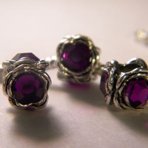 May include: Three silver charm beads with purple gemstone accents. Each bead features a decorative floral design.