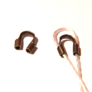 Puede incluir: Dos cuentas de crimpado de metal dorado, una en un cordón blanco, utilizadas para la elaboración de joyas.