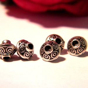 May include: Close-up of six silver-colored beads with intricate circular and swirl patterns. The beads have a metallic sheen and are designed with a hole for stringing. The beads are likely for jewelry making.