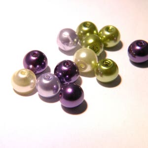May include: A collection of round, pearl-like beads in shades of purple, green, and cream. The beads are arranged on a white surface.
