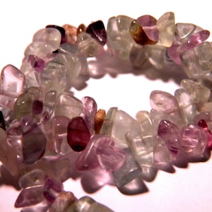 Peut inclure: Un collier de perles de pierres précieuses roses, violettes et blanches. Les perles sont de formes et de tailles irrégulières et sont enfilées sur un fil transparent.
