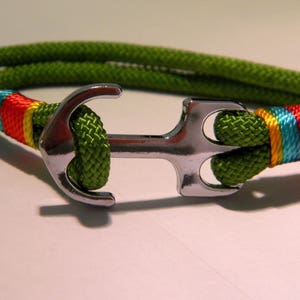 Puede incluir: Un brazalete de cuerda verde con un dije de ancla de plata. La cuerda está acentuada con una franja multicolor de azul, amarillo, naranja y rojo.