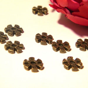 May include: A collection of ten bronze-colored metal flower-shaped charms. The charms are scattered on a white surface.