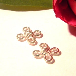 Könnte beinhalten: Zwei silberfarbene Metallverschlüsse, jeweils mit einem Design aus vier Schlaufen, werden vor einem weichen, cremefarbenen Hintergrund gezeigt. Die Verschlüsse haben eine dekorative, symmetrische Form und werden wahrscheinlich für Schmuck oder Bastelprojekte verwendet.