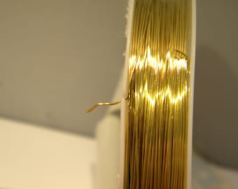 Pode incluir: Um carretel de fio de cor dourada, enrolado firmemente em torno de um núcleo de plástico branco. O fio é fino e brilhante, e a ponta do fio é visível, estendendo-se do carretel.