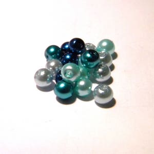 Peut inclure: Un assortiment de perles de couleur bleu turquoise, bleu et blanc. Les perles sont disposées en un tas lâche sur une surface blanche.