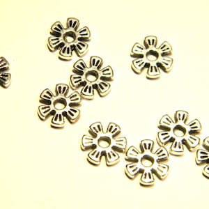 以下が含まれることがあります： 中央に穴が開いた銀色の金属製のフラワー型ビーズが10個あります。ビーズは白い背景に散らばって配置されています。