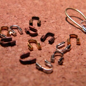Puede incluir: Una colección de pequeños cierres de metal en forma de bucle en tonos plateado, dorado y cobre. Los cierres están dispuestos sobre una superficie de corcho.
