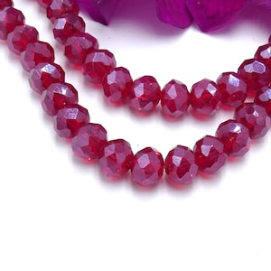 May include: Close-up of two strands of faceted, deep red beads. The beads have a sparkling, gem-like quality, reflecting light. The background is white with a hint of purple. These beads are suitable for jewelry making.