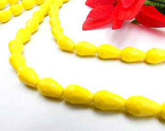 May include: A close-up of a yellow beaded necklace with a teardrop shape. The necklace is displayed on a white surface, with a red flower in the background. The beads are evenly spaced and have a glossy finish.