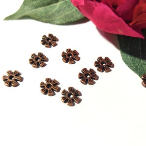 May include: A collection of eight copper-colored metal flower-shaped beads. The beads have a detailed design and are arranged on a white surface.